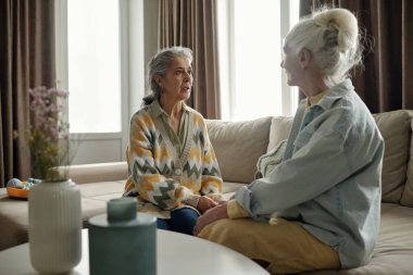 Oturma odasında oturan iki yaşlı kadın, pencerelerden içeri doğal ışık süzülen rahat bir dekorla çevrili bir şekilde sohbet ediyorlar. Sahne sıcaklık ve bağlantı öneriyor