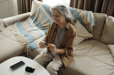 Oturma odasındaki kanepede otururken akıllı telefona bakan son sınıf kadını. Arka planda mavi çizgili bir battaniye ve kablosuz hoparlörlü beyaz bir sehpa var.