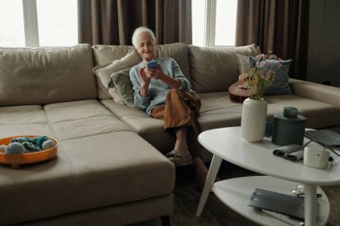 Modern oturma odasında elinde bir cep telefonu, yanında da gitar ve vazoyla koltukta rahat bir şekilde oturan yaşlı bir insan.