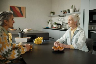 Mutfak masasında oturmuş kahvaltıda sohbet eden iki son sınıf öğrencisi. Çörek, muz ve fincanlı masa sıcak, sade bir atmosfer yaratır.