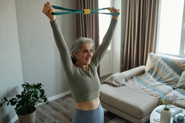 Hafif atletik giyimli yaşlı kadın oturma odasında direniş bandıyla esniyor, parlak doğal ışık ile çevrili, halinden memnun ve odaklanmış görünüyor.