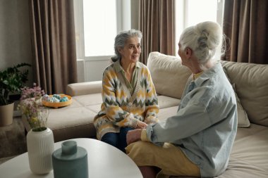 İki yaşlı kadın oturma odasındaki kanepede oturmuş, içtenlikle sohbet ediyorlar, etraflarında bitkiler ve yumuşak mobilyalar var.