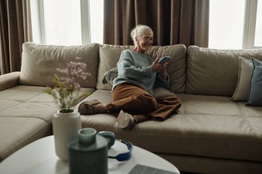 Yaşlı bir kadın bej renkli rahat bir kanepeye oturmuş elinde akıllı bir telefonla gülümsüyor. Oturma odasında pencerelerden süzülen doğal ışık ve masada vazo.