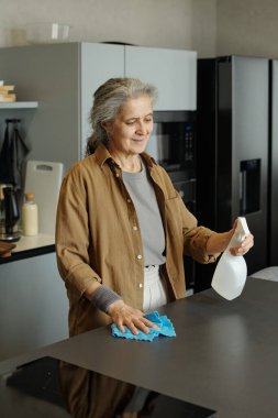 Kahverengi tişört giyen yaşlı kadın elinde sprey şişeyle gülümsüyor ve mutfak tezgahını mavi bezle temizliyor.