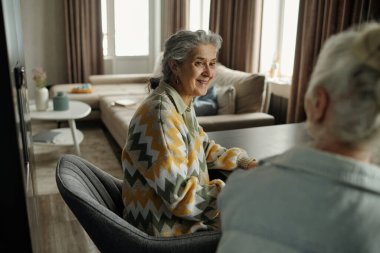 Yaşlı kadınlar, İskandinav tasarımı ile samimi, modern oturma odasında dostça sohbet ediyorlar. Arkaplanda yumuşak ışıklandırma, çağdaş mobilyalar ve geniş pencereler yer alıyor