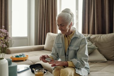 Kanepede oturan yaşlı bir kadının portresi, tıbbi aletler kullanmaya odaklanıyor. Etrafında çiçekler, çay fincanı ve dekoratif tepsi bulunmakta olup, rahat bir atmosfer oluşturmaktadır.