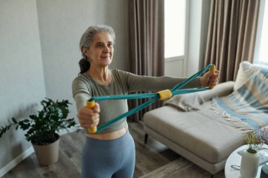 Modern oturma odasında direniş bandıyla egzersiz yapan yaşlı bir kadın aktif bir yaşam tarzı sergiliyor. Bitki ve rahat kanepe arka planda görünür