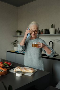 Tezgahta ekmek, meyve ve sebzelerle modern bir mutfakta telefonla konuşurken elinde çay tutan yaşlı bir kadın. Arka planda çeşitli bitki ve mutfak gereçleri görünür.