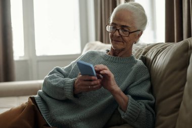 Gözlüklü yaşlı kadın akıllı telefonuyla uğraşırken rahat koltuğun keyfini çıkarıyor. Rahat oturma odasında rahatlatıcı ve sakin bir atmosfer. Pencereden içeri ışık akıyor.
