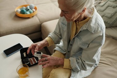 Tansiyonunu dijital cihazla ölçen yaşlı bir kadın, arka planda çay ve ilaç organizatörüyle kanepede otururken...