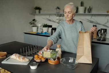 Mutfak tezgahına erzak paketini boşaltan yaşlı bir kadın yemek malzemeleri hazırlıyor. Ekmek, yumurta ve sebze içerir, arka planda modern mutfak aletleri görünür.