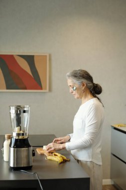 Modern mutfakta duran yaşlı bir kadın, malzemeleri mikserle karıştırıyor. Taze muzlar ve kesme tahtası tezgahın üstünde ve arka planda soyut sanat eserleri asılı.