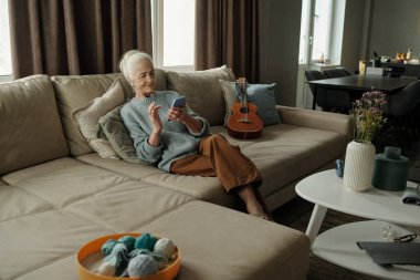 Kanepede oturan, iplik ve iğnelerle örgü ören yaşlı bir kadın. Yan koltukta dinlenen gitar ve modern mobilyaları olan rahat, aydınlık bir oda.