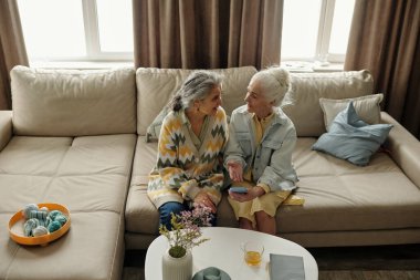Yaşlı kadınlar bej bir koltukta oturup sohbet ediyorlar. Kahve fincanları ve dekoratif malzemelerle bir masanın etrafında oturuyorlar.