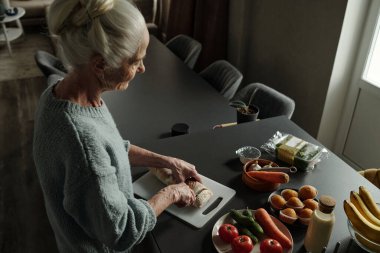 Çevresinde taze sebzelerle ekmek dilimleyen yaşlı bir kadın, huzurlu bir mutfak ortamında yemek pişirme faaliyetlerine girişiyor.