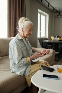 Oturma odasında otururken bir glukometre cihazıyla meşgul olan yaşlı bir kadın. Öğleden sonra ışığı usulca etrafını aydınlatıyor.