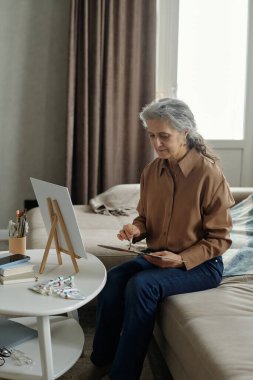 Oturma odasında rahat bir şekilde resim yapmaya odaklanmış yaşlı bir kadın yanında sanat malzemeleri ve pencereden içeri giren doğal ışıkla oturuyordu.