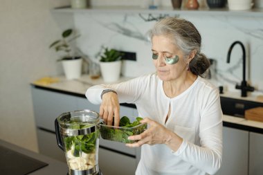 Smoothie hazırlığı için modern mutfakta taze ıspanak ve muzları harmanlayan, sağlıklı yaşam tarzı ve kişisel bakım rutinini gösteren, göz altı yamaları olan yaşlı bir kadın.