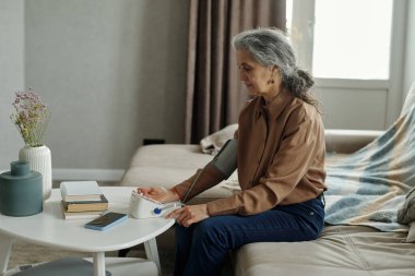 Kanepede oturan yaşlı bir kadın dijital bir aletle kan basıncını ölçüyor. Modern oturma odası, vazodaki çiçekler ve masadaki kitaplar rahatlatıcı bir atmosfer yaratıyor.