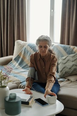 Rahat bir koltukta otururken kan basıncı monitörü kullanan yaşlı beyaz bir kadının portresi. Yanında akıllı telefon ve fincanla odaklanmış ve özenli görünüyor.