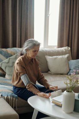 Yastıklar ve tıbbi malzemeler de dahil olmak üzere rahat bir çevresi olan kanepede otururken tansiyonunu ölçen günlük kıyafetli yaşlı bir kadın.