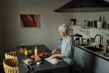 Modern mutfak ortamında taze sebzelerle ve malzemelerle mutfak tezgahının etrafında çeşitli mutfak aletleriyle yemek hazırlayan yaşlı bir kadın.
