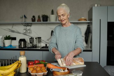 Son sınıf öğrencisi kadın, modern mutfakta çeşitli taze sebze ve yumurtalarla ekmek kesiyor. Aşçılık faaliyetlerine ilgi duyuyor gibi görünüyor.