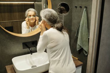 Modern banyo aynasında duran son sınıf öğrencisi kadın, gözlükleri ayarlıyor. Ahşap elementlerle çağdaş bir ortamda yakalanmış.