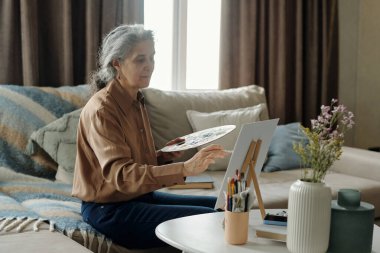 Oturma odasındaki masada oturan, etrafı mobilya ve sanat malzemeleriyle çevrili, yaratıcı ifade ve rahatlamaya odaklanmış son sınıf öğrencisi bir kadın.