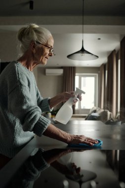 Modern bir evde mutfak tezgahını temizlerken elinde sprey şişesi tutan son sınıf kadını. Ev temizliğini korumak için kendini adamış ve çaba sarf etmiş.