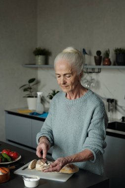 Üst düzey kadın, çeşitli sebzeler ve kesim tahtasıyla donatılmış modern mutfakta yemek hazırlarken ekmek dilimlemeye odaklanmış.