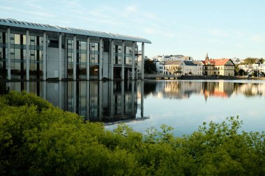 Güneşli bir günde, nehir kıyısındaki eski kasabanın yakınındaki yeşil çalılıklara karşı panoramik pencereleri ve beton sütunları olan modern bir bina manzarası.