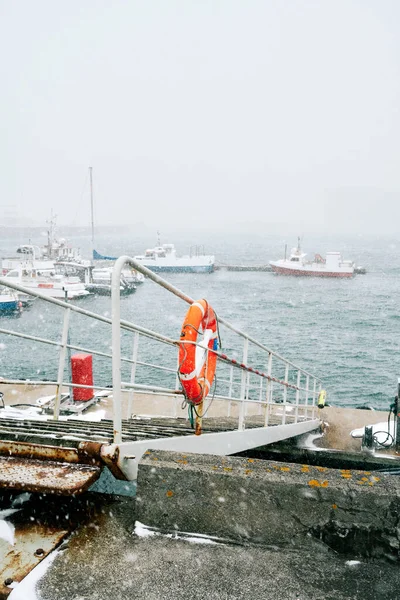 冬季用小船在通往靠近波涛汹涌大海的港口的人行道栏杆上的救生圈 — 图库照片