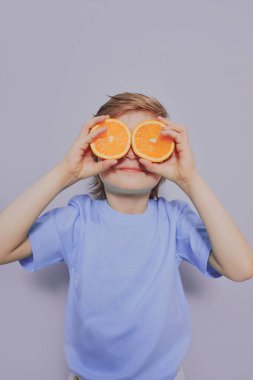 Beyaz tişörtlü kıvırcık Afrikalı Amerikalı çocuk ellerinde iki portakal tutuyor ve gri bir arka planda gülümsüyor..