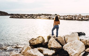 Gün batımında denizin önünde taşların üzerinde bir kadın