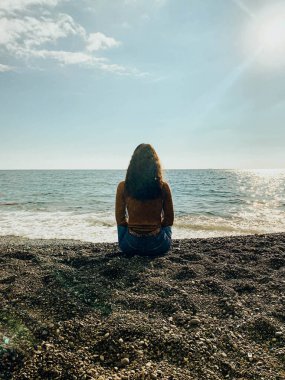 Siyah taş plajda oturan kadın denize bakıyor. Güneşe bakıyor.