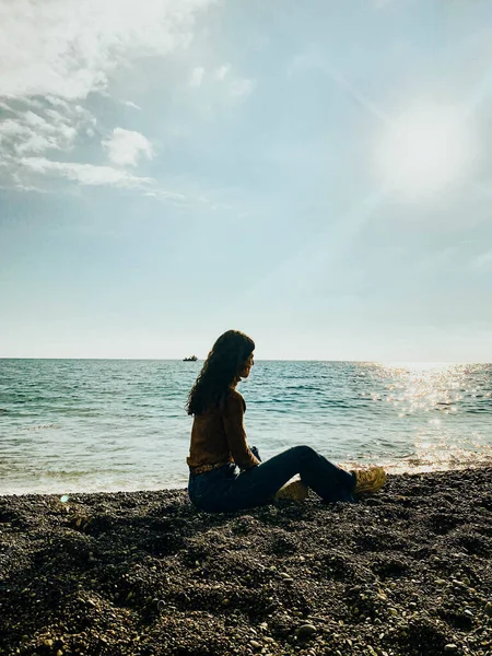 海に面した黒い石のビーチに座って太陽を見ている女性は — ストック写真