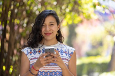 Real Mexican woman holding smart phone and looking at camera outdoors