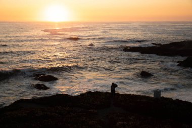 Arka planda genç bir kadın okyanusun üzerinde günbatımını fotoğraflıyor.