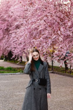 Yünlü ceketli genç bir kadın baharda kiraz çiçeklerinin arasında yürür.