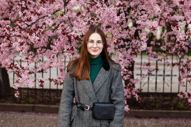 Yünlü ceketli genç bir kadın baharda kiraz çiçeklerinin arasında yürür.