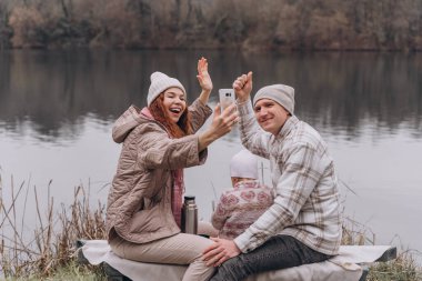 Üç kişilik bir aile sonbahar parkında nehir kenarında oturup selfie çekiyor.