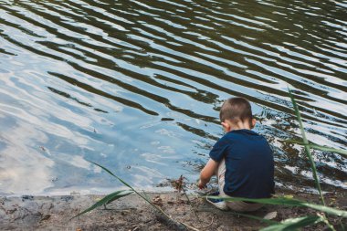 Küçük çocuk nehir kıyısının arka manzarasına çömeliyor.