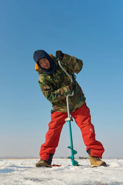 暖かい服を着たアジアの男が冬の釣りで氷の穴を開ける — ストック写真