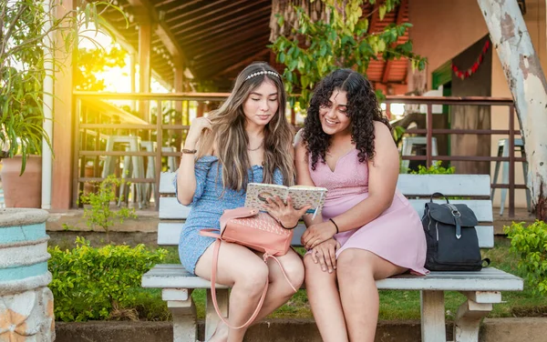 Two smiling friends sitting reading the same book on a bench outdoors