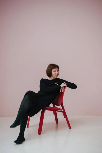 穿着黑色连衣裙的女人坐在椅子上 粉色背景 — 图库照片