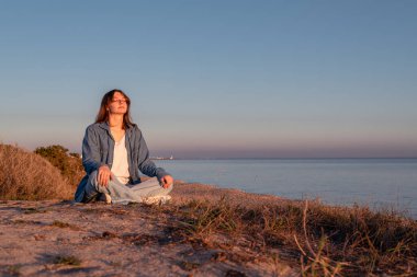 Kotlu genç bir kadın gün batımında deniz kıyısında meditasyon yapıyor.