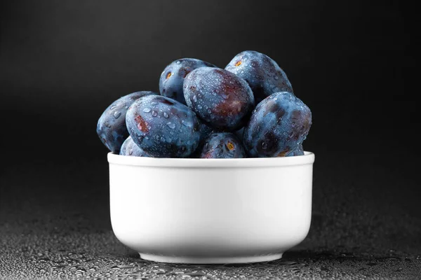 Blue plums in a white plate with water drops on a dark background. Useful dietary fruit.