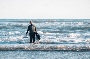 Dalgıç kıyafetli genç elinde sörf tahtasıyla sörf tahtasına doğru yürüyor.