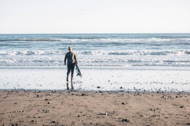 Dalgıç kıyafetli genç elinde sörf tahtasıyla suya giriyor.